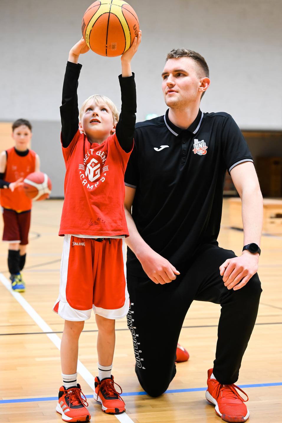 Ein junger Basketballspieler hebt den Ball über seinen Kopf, während ein Trainer neben ihm kniet und zuschaut.