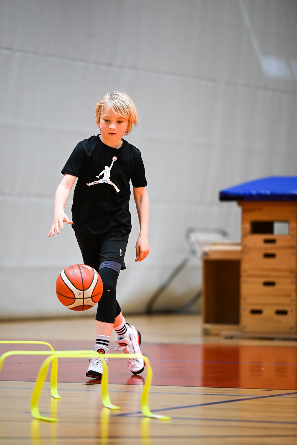 Junge dribbelt einen Basketball über gelbe Hürden in einer Sporthalle. Er trägt ein schwarzes T-Shirt und Sporthose.