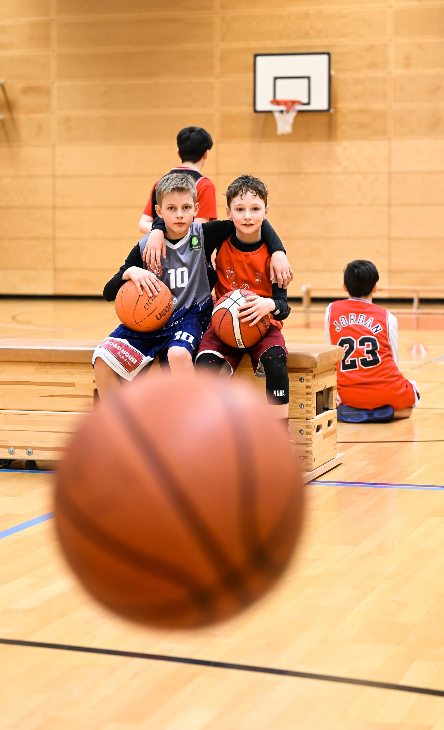 Zwei Jungen sitzen auf einer Bank in einer Sporthalle, jeder mit einem Basketball in der Hand.