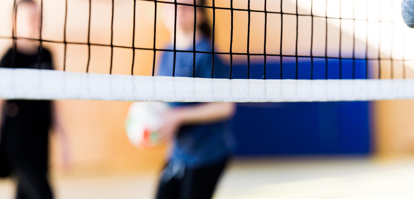 Unscharfe Silhouetten von zwei Personen hinter einem Volleyballnetz in einer Sporthalle. Eine Person hält einen Volleyball.